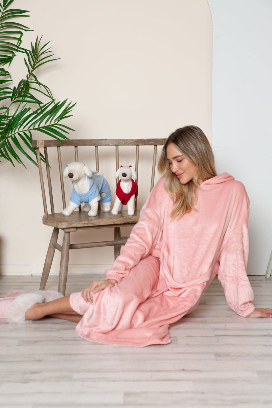 Full size pocketed hooded midi lounge dress in pink worn by a woman sitting on a floor with plush dog toys on a chair in the background.