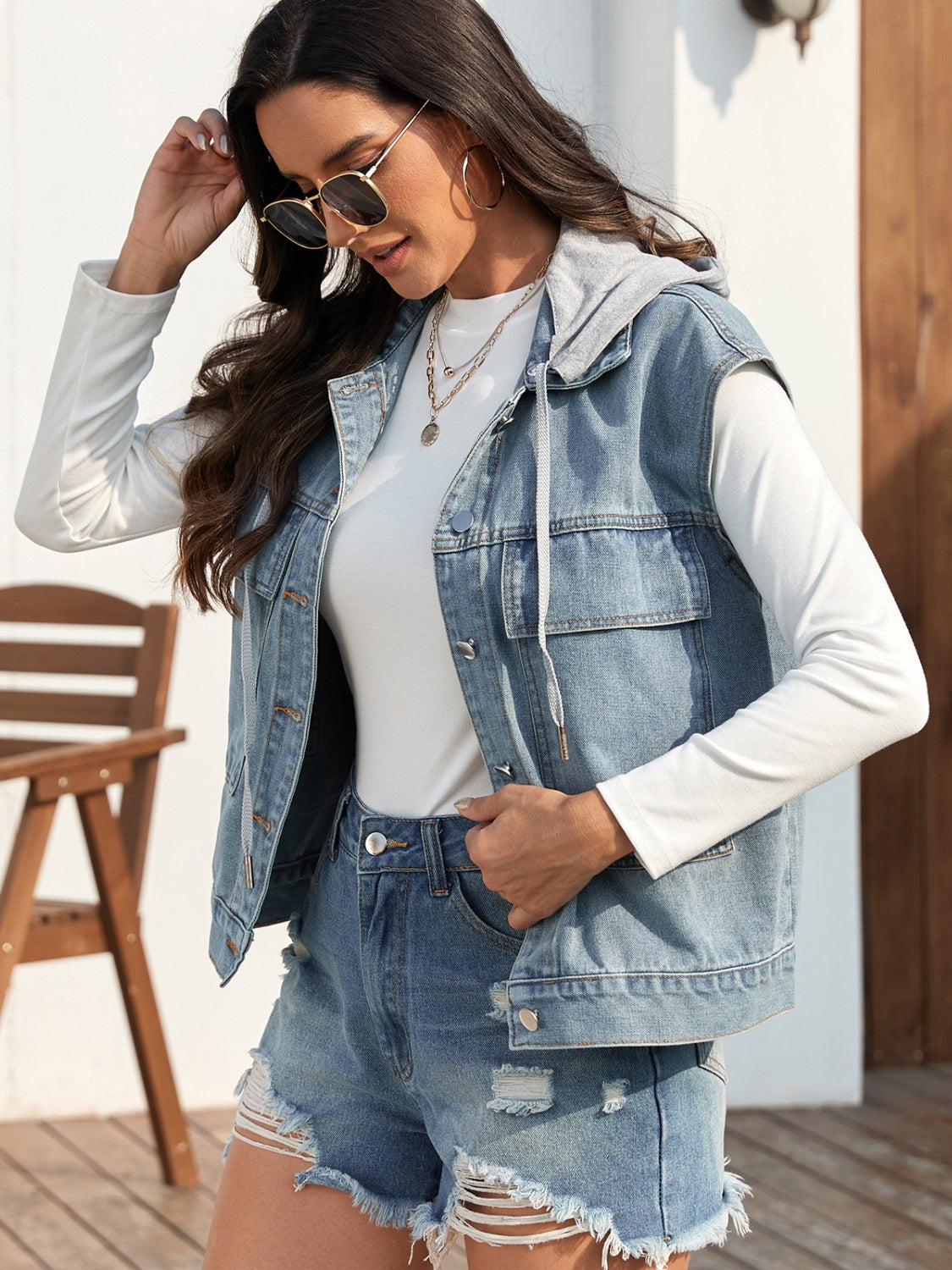 Sleeveless denim jacket with drawstring and button-up design, worn over a white longsleeve top.