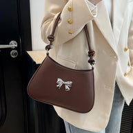 Small bow PU leather knotted strap handbag in brown displayed on shoulder.